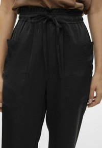 Zwarte broek met een elastische tailleband, strik vooraan en twee zijzakken. De stof heeft een gladde textuur en een relaxte pasvorm.