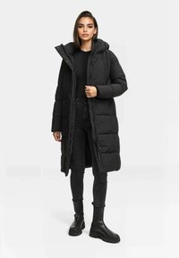 Abrigo de plumas negro largo con capucha, botones a presión y diseño acolchado, llevado sobre una camiseta negra y jeans ajustados, combinado con botas negras.
