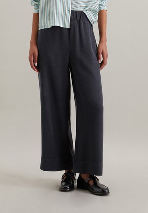 Wijdvallende, donkergrijze broeken van zacht materiaal met een elastische taille. Gecombineerd met een gestreepte blouse en zwarte schoenen.