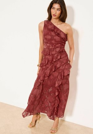 Kvinde iført en burgunderrød, blomsterprydet, asymmetrisk kjole med flæser og gyldne høje hæle, stående mod en ensfarvet væg.