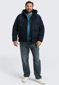 Gewatteerde marineblauwe jas met een rits aan de voorkant, verstelbare capuchon en vier zakken, gedragen over een blauwe shirt en gecombineerd met lichtblauwe jeans en witte schoenen.