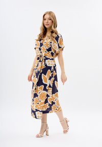 Robe marine à imprimé floral avec manches courtes, taille cintrée et jupe fluide. Agrémentée de fleurs orange et de tourbillons. Associée à des talons à lanières.