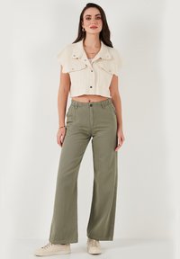 Kurzes cremefarbenes Hemdjackett mit Schulterklappen, kombiniert mit lockeren olivfarbenen Hosen. Model trägt beige Turnschuhe.