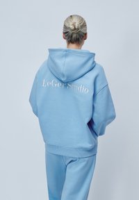 Hanorac albastru deschis cu o capotă mare, croială supradimensionată și tiv cu nervuri. Are branding brodat alb pe spate. Este asortat cu pantaloni corespunzători.