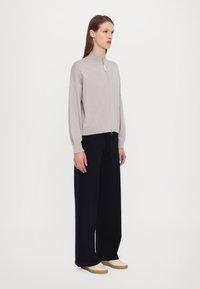 Ljusgrå zip-up-tröja med en ståkrage, lösa ärmar och elastisk trim, kombinerad med breda svarta byxor och beigefärgade skor.
