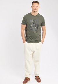 T-shirt en coton vert olive avec un imprimé graphique comportant des rayures et un design de disque. Assorti à un pantalon en velours côtelé crème et des chaussures bateau marron.