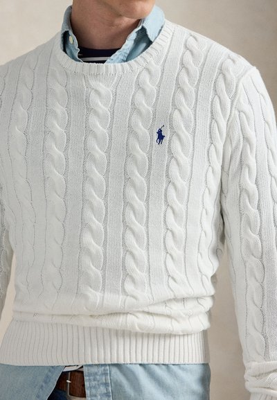Pull en maille torsadée blanche avec un logo bleu brodé, porté par-dessus une chemise bleu clair à boutons et un t-shirt rayé.
