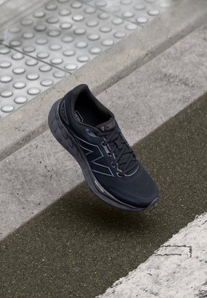 Zwarte sportschoen met "N"-logo en dikke zool, zwevend boven een getextureerde stoeprand en een asfaltweg met witte lijn.