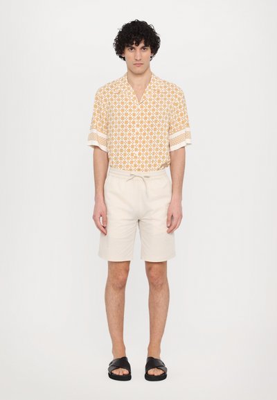 Chemise à manches courtes à motif géométrique orange et crème, assortie à un short beige clair. Des sandales noires complètent la tenue.