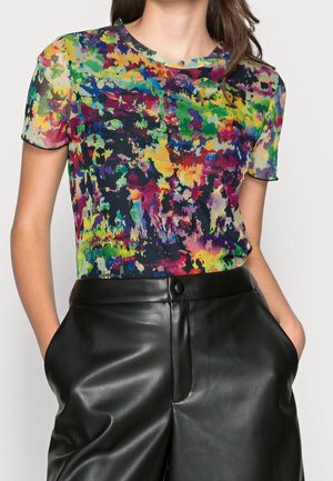 Femme portant un haut à manches courtes avec un imprimé abstrait multicolore et un pantalon en cuir noir taille haute, les mains dans les poches.