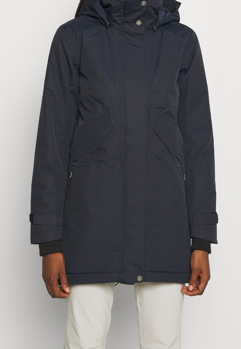 Personne portant un manteau d'hiver à capuche bleu marine foncé avec fermeture éclair et boutons-pression, poches à rabat et pantalon beige, debout devant un fond clair.