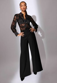 Crna čipkasta košulja sa satenskim detaljima, uska u struku, uparena s crnim širokim hlačama visokog struka i crnim cipelama s oštrim vrhom.