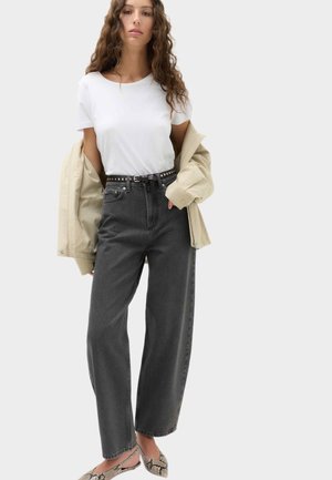 Kvinde med krøllet hår iført hvid t-shirt, beige jakke hængende over armene, sort højtaljede jeans med søm-bælte og flade sko med slangeskind.