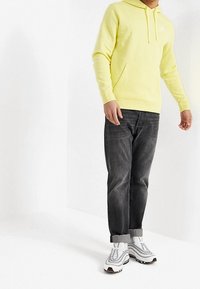 Pánská neónově žlutá mikina s kapucí a přední kapsou, v kombinaci s tmavými džíny a bílými teniskami s černými prvky.