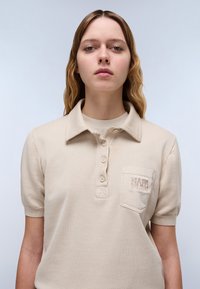 Vaaleanbeige polo-paita, jossa on kaulus, kahden napin halkio, lyhyet hihat ja rintatasku, jossa on saman värinen brodeerattu logo-otsikko.