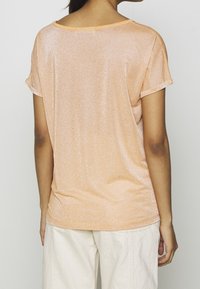 Lyhythihainen, kimalteleva persikkatoppi, jossa on rento malli ja pyöreä kaula-aukko, joka näyttää sileältä ja hienovaraisesti kiiltävältä.