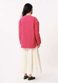 Veste rose matelassée avec des manches longues, portée avec une jupe blanche à volants et des chaussures plates vertes. Le tissu a un motif texturé.