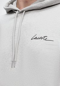 Sweat à capuche gris clair avec un logo Lacoste noir sur la poitrine gauche, avec des cordons suspendus à la capuche.