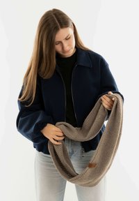 Marineblauwe ritsjack met opgeblazen mouwen, gedragen over een zwarte trui, gecombineerd met een gestructureerde beige gebreide sjaal die in de handen wordt gehouden.