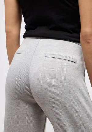 Close-up van een persoon die lichtgrijze broek met visgraatpatroon draagt, voorzien van achterste inschuifzakken, en een zwart mouwloos topje.