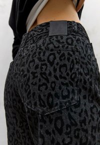 Črne jeanske hlače iz teksturiranega leoparda, ki vključujejo zadnji žep in črn usnjen značko na pasu.