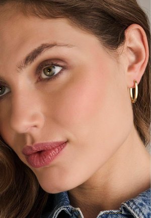 Guld hoops ørering med en glat, poleret overflade, der har en cirkulær form og et slankt design. Nærbillede mod hudfarve.