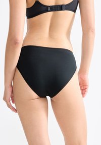 Vrouw in zwart hoogoplopende ondergoed en bijpassende beha, van achteren gezien tegen een witte achtergrond.