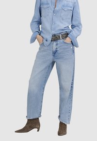 Jeans en denim bleu clair avec une coupe décontractée, présentant des détails usés. Associés à une chemise boutonnée bleu clair et des bottines en suède marron.