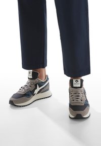 Sneakers sportivi con una combinazione di grigio, blu navy e bianco, caratterizzati da un design in suede e rete, colletto imbottito e suola in gomma testurizzata.