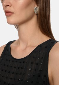 Schwarzes ärmelloses Oberteil aus einem transparenten Stoff mit einem Gittermuster, akzentuiert durch kleine schwarze Pailletten. Das Model trägt einen großen silbernen Ohrring.