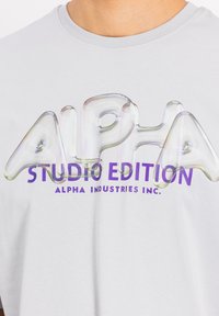 Siva majica s glatkom teksturom koja ima iridescentni natpis "ALPHA" i ljubičasti print "STUDIO EDITION". Ležeran okrugli izrez.