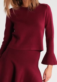 Bordowa, długorękawowa bluzka z okrągłym dekoltem i rozkloszowanymi rękawami, wykonana z gładkiej tkaniny. Charakteryzuje się krótkim krojem i minimalnymi przeszyciami.