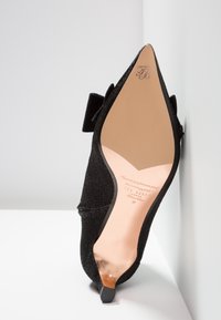 Talons noirs pointus avec une tige en tissu texturé, ornés d'un accent de nœud. La semelle est rose avec un logo, mettant en avant une silhouette élancée.