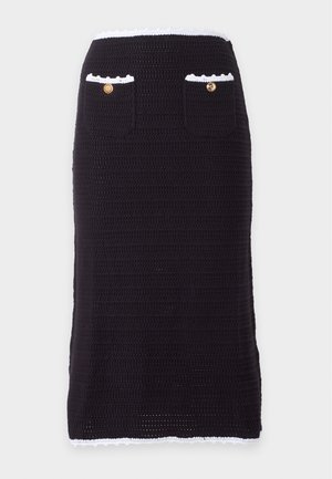 Femme portant un gilet noir sans manches boutonné et une jupe assortie avec un liseré blanc, tenant un grand sac cabas tissé beige.