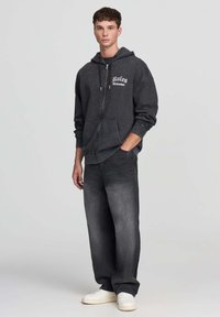 Giovane in piedi con una mano in tasca, indossando una felpa nera con zip con la scritta "Rules Chinatown", jeans scuri e sneakers bianche.