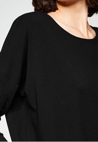 Svart stickad topp med vid, rund halsringning och lös passform. Mjuk textur med sänkta axlar. Sömdetaljer längs sidorna.