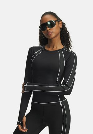 Zwart lange mouwen sporttop met witte accentnaden en meshpanelen, gecombineerd met zwarte leggings en futuristische zwarte zonnebril.