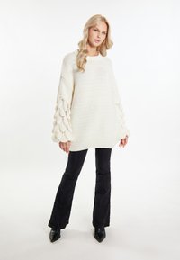 Pulover gros cu tricotare crema, cu manșete texturate și ondulate; asortat cu pantaloni negri evazați și cizme negre cu vârful ascuțit.