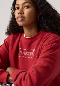 Červená flísová mikina s bílou vyšívanou klasikou "Levi's" na přední straně. Materiál je měkký a má design s kulatým výstřihem.