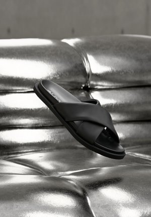 Svart skinn glide-sandal med kryssede stropper vist over en blank sølvfarget polstret overflate.
