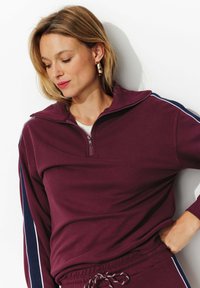 Sweatshirt zippé bordeaux avec col montant, doté de bandes latérales bleu marine et blanches. Tissu doux avec une coupe décontractée et des poignets côtelés.