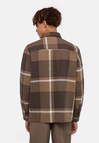 Camicia a quadri marrone con grandi quadrati in marrone scuro, beige e bordeaux. Presenta un colletto con bottoni e maniche lunghe con polsini abbottonati.
