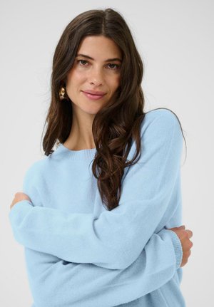 Junge Frau mit langen dunklen Haaren, die goldene Creolen und einen hellblauen Pullover trägt, mit verschränkten Armen und sanft lächelnd vor einem neutralen Hintergrund.