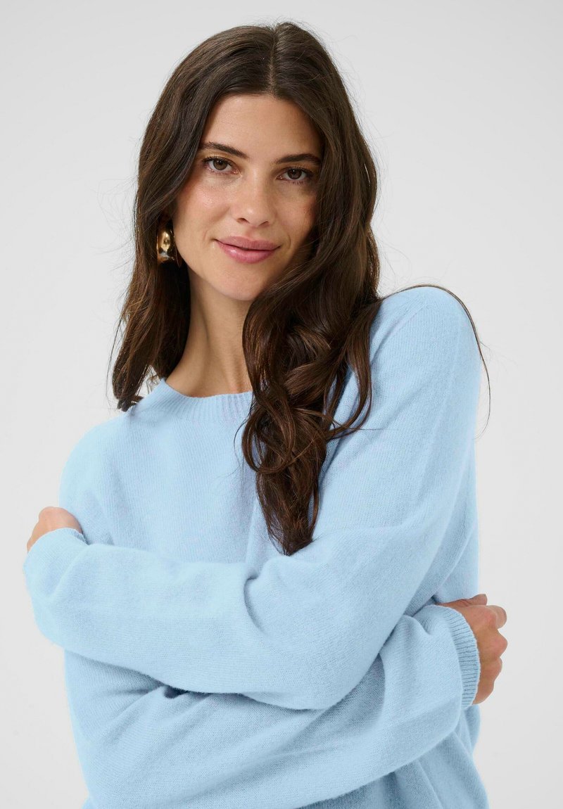 Fiatal nő hosszú sötét hajjal, arany fülbevalókkal és világoskék pulóverben, karba tett kézzel, lágyan mosolyogva egy semleges háttér előtt.