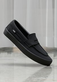 Svart lær slip-on sko med rund tå, teksturert overflate og frontklaff. Fungerer med Vans-logo på siden.