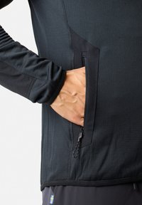 Zwart sporttop met een veiligheidszak met rits, lange mouwen en een textuurstof met platte naden aan de zijkanten.