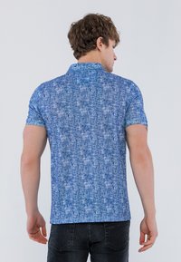 Kortærmet blå polo shirt med et tekstureret mønster, krave og lige kant. Bæres med mørke jeans, vist fra ryggen.