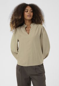 Beige blúz V-nyakkal és fodros gallérral, hosszú puffos ujjakkal és laza fazonnal. Az anyaga puha, texturált felületű.