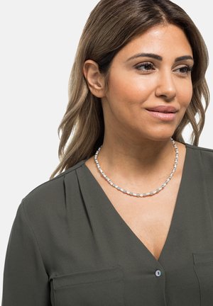 Silberne Perlenkette, die von einem Modell getragen wird, mit ovalen Perlen in glänzender Optik, vor einem dunklen grünen Oberteil mit V-Ausschnitt.