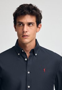 Dunkelblaues Hemd mit Knopfleiste, aus glattem Stoff; verfügt über weiße Knöpfe und ein kleines rotes Logo, das eine Figur mit einem Golfschläger zeigt.